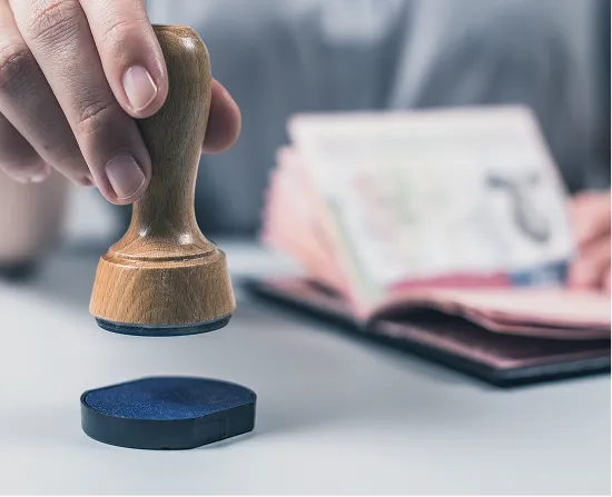 A close-up of a hand holding a wooden stamp above an ink pad, with an open passport blurred in the background