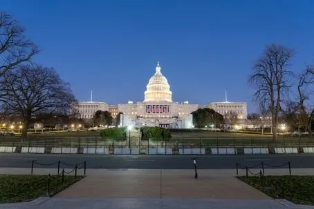 A wide view of a large domed government building illuminated at dusk, with lights glowing against a clear blue sky and bare trees surrounding the foreground.