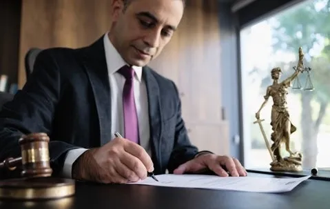 A lawyer signs a document, beside him are a wooden gavel and a justice scale.