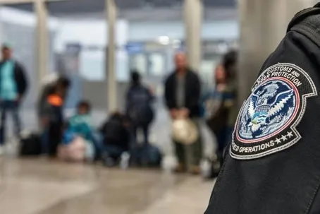 A close-up of a U.S. Customs and Border Protection officer’s uniform patch, with a blurred group of people standing and sitting in the background inside an immigration or security area