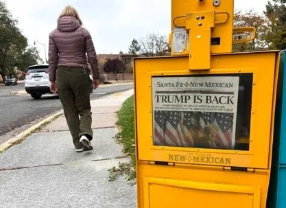A yellow newspaper vending box on a sidewalk displaying a front page with the headline ‘Trump Is Back,’ while a person walks away in the background on a suburban street.